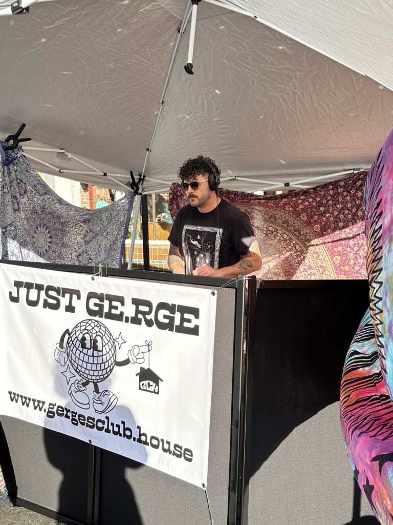 just ge.rge DJing at the san luis obispo farmer;s market. He is standing under a white pop up tent, behind a black dj facade with a sign that says just ge.rge and has his logo